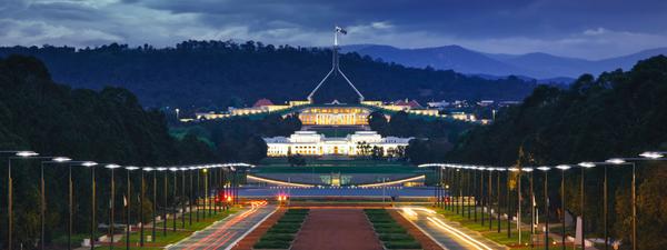 Evening view of Australian Parliament House in Canberra, ACT, with illuminated facade and flag, surrounded by empty streets and trees.