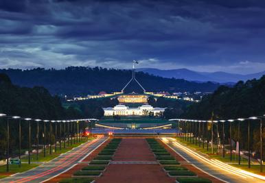 Evening view of Australian Parliament House in Canberra, ACT, with illuminated facade and flag, surrounded by empty streets and trees.